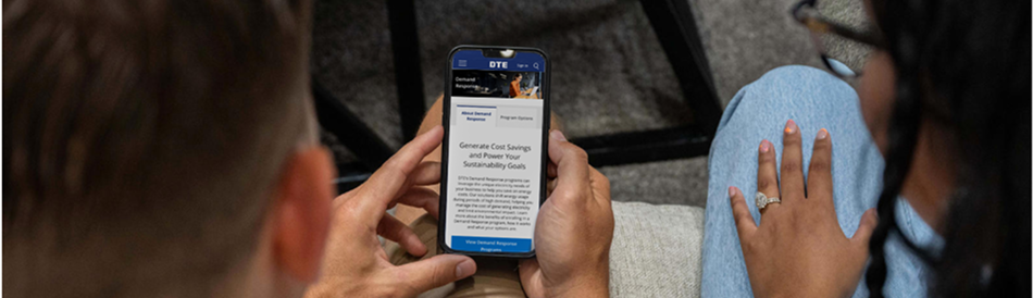 A person is holding a smartphone displaying a webpage from DTE Energy about generating cost savings and achieving sustainability goals. Another individual is seated nearby, partially visible. The setting appears to be indoors with a casual environment, and the phone screen shows program details related to energy efficiency.